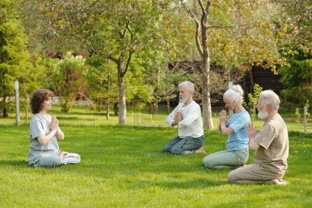 Téléchargez les photos : Vue de côté de la jeune enseignante de yoga et du groupe de patients âgés de la maison de retraite pratiquant l'exercice de méditation sur la pelouse verte - en image libre de droit