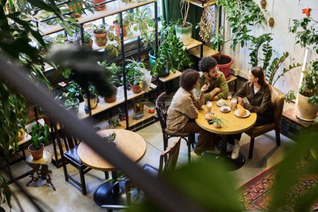 Téléchargez les photos : Groupe de jeunes gens interculturels réunis par table ronde en bois dans un café confortable prendre un café et des collations tout en discutant des dernières nouvelles - en image libre de droit