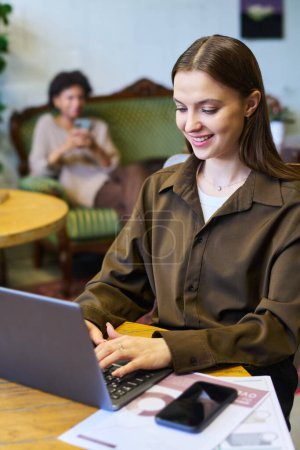 Foto de Joven mujer de negocios moderna en ropa de casualwear inteligente escribiendo en el teclado del ordenador portátil mientras se sienta junto a la mesa en la cafetería contra la compañera de trabajo - Imagen libre de derechos
