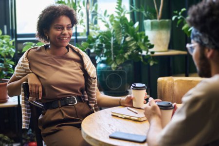 Téléchargez les photos : Jeune femme afro-américaine avec une tasse de café assise à table devant ses amis et regardant l'un d'eux pendant la conversation - en image libre de droit