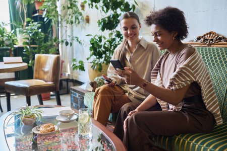 Foto de Dos novias interculturales sonrientes viendo videos divertidos en línea en un teléfono inteligente mientras están sentadas en un cómodo y elegante sofá en la cafetería - Imagen libre de derechos