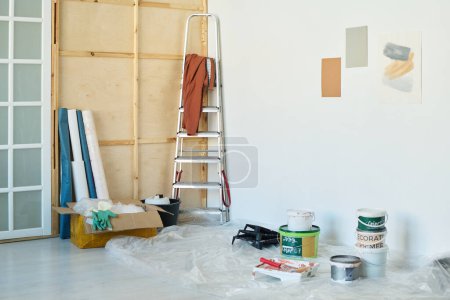 Photo for Corner of spacious living room of new apartment with buckets of paints or glue for wallpapers, stepladder, box with rubber gloves and other stuff - Royalty Free Image