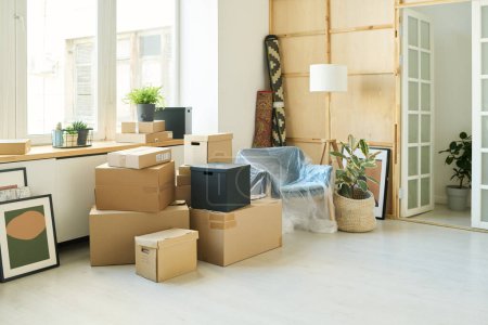 Stack of boxes, pictures, green domestic plants and armchair standing by large window of living room of new apartment during relocation
