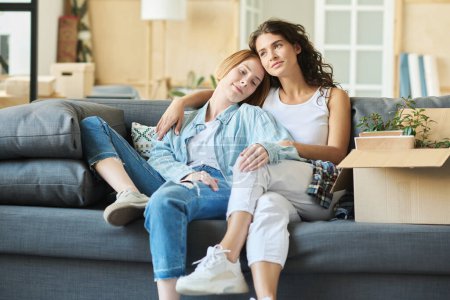 Téléchargez les photos : Deux jeunes copines heureuses en tenue décontractée assises sur un canapé confortable dans le salon et profitant du repos après leur déménagement dans un nouvel appartement - en image libre de droit
