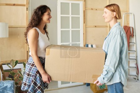Photo for Side view of two happy girls in casualwear carrying big cardboard box and looking at one another during relocation to new apartment or house - Royalty Free Image