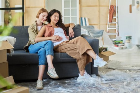 Foto de Joven mujer rubia abrazando a su novia y tocando su barriga embarazada mientras ambos están sentados en un cómodo sofá en su nuevo apartamento - Imagen libre de derechos