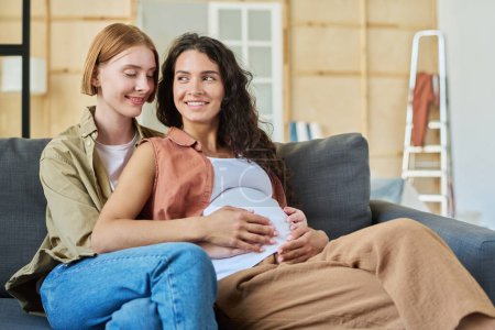 Photo for Two happy young women sitting on couch in front of camera after relocation while one of them embracing her pregnant girlfriend - Royalty Free Image