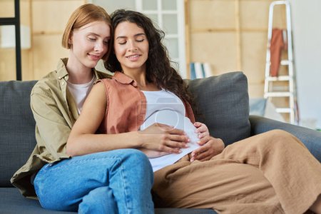 Photo for Happy young brunette pregnant woman and her affectionate girlfriend sitting on couch and enjoying rest after relocation to new apartment - Royalty Free Image