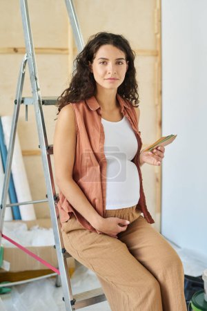 Foto de Joven mujer morena embarazada con paleta de colores en la mano mirando a la cámara mientras se sienta en la escalera y elegir la pintura para las paredes - Imagen libre de derechos