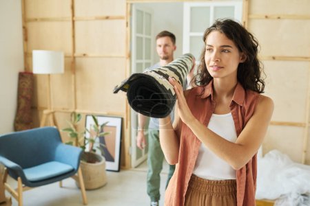 Photo for Young brunette woman holding rolled carpet over shoulder while carrying it with her husband or boyfriend during relocation to new apartment - Royalty Free Image