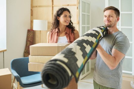 Photo for Happy young man holding rolled carpet on his shoulder while moving forwards along living room with his wife and carrying it during relocation - Royalty Free Image