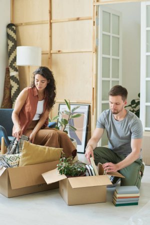 Photo for Young man and woman packing cardboard boxes with books, green domestic plants and bedclothes while moving for new apartment - Royalty Free Image