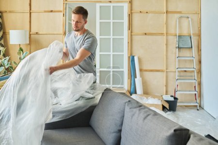 Photo for Happy young man in casualwear covering new couch with cellophane packaging while preparing spacious living room for renovation - Royalty Free Image