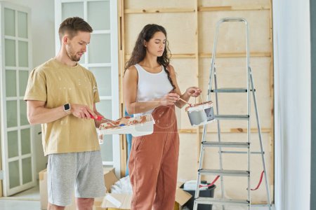 Photo for Young couple in casualwear holding plastic bucket and tray while mixing colors before painting walls of spacious living room of new apartment - Royalty Free Image