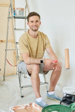 Photo for Young smiling man with paintroller sitting on stepladder by white wall of living room and looking at camera during renovation of apartment - Royalty Free Image
