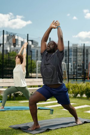 Téléchargez les photos : Jeune homme actif afro-américain en vêtements de sport debout sur le tapis sur le terrain de sport et l'exercice tout en gardant les bras levés au-dessus de la tête - en image libre de droit