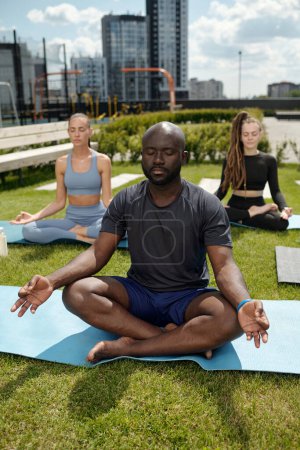 Foto de Joven hombre afroamericano sereno en camiseta y pantalones cortos practicando yoga mientras está sentado en la esterilla en pose de loto contra dos mujeres en chándales - Imagen libre de derechos