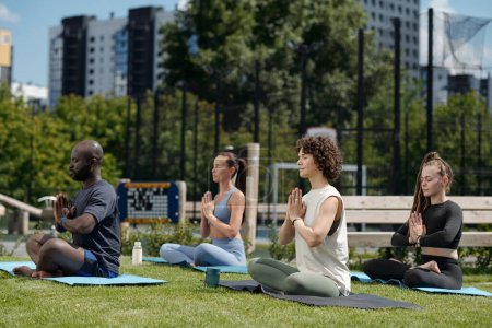 Photo for Side view of group of intercultural athletes practicing yoga outdoor while sitting on mats with their legs crossed and palms put together by chest - Royalty Free Image