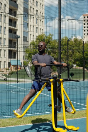 Foto de Joven deportista afroamericano haciendo ejercicio en instalaciones deportivas mientras entrena sus piernas contra el campo de fútbol y edificios modernos - Imagen libre de derechos