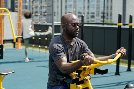 Téléchargez les photos : Homme afro-américain actif avec des lunettes et un t-shirt pompant les muscles des bras sur les installations sportives devant la caméra contre la sportive - en image libre de droit