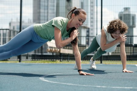 Two young active female athletes doing difficult physical exercise on sports ground on sunny summer day against buildings in urban environment