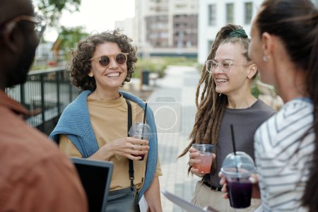 Foto de Dos jóvenes riendo mujeres caucásicas con refrescos mirando a sus colegas interculturales mientras charlan en el descanso en el entorno urbano - Imagen libre de derechos