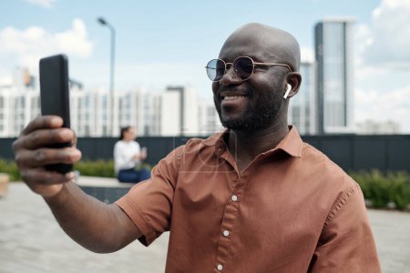 Téléchargez les photos : Jeune homme afro-américain joyeux dans les écouteurs regardant l'écran du smartphone tout en communiquant avec quelqu'un dans le chat vidéo en plein air - en image libre de droit