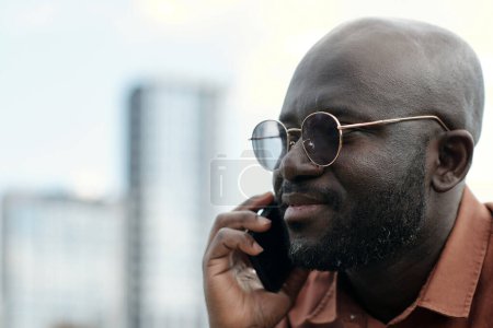 Photo for Close-up of young confident African American businessman in eyeglasses holding smartphone by ear while talking to someone outdoor - Royalty Free Image