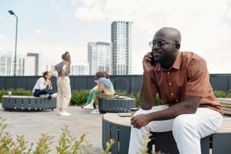 Téléchargez les photos : Jeune homme d'affaires confiant avec smartphone par l'oreille assis sur le banc dans le parc et de communiquer avec quelqu'un contre un groupe de femmes - en image libre de droit