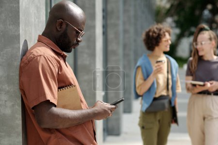 Téléchargez les photos : Jeune homme d'affaires afro-américain sérieux textos dans un téléphone mobile sur une journée ensoleillée d'été tout en se tenant près du mur gris du bâtiment moderne - en image libre de droit