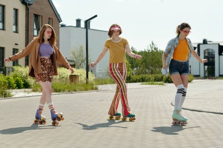 Téléchargez les photos : Trois adolescentes actives en tenue décontractée patinant sur roulettes en milieu urbain et profitant de loisirs le jour ou le week-end ensoleillés d'été - en image libre de droit