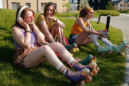Téléchargez les photos : Jeune femme avec écouteurs écouter de la musique tout en étant assis sur la pelouse verte et se reposer avec ses amis après le patinage à roulettes dans la ville - en image libre de droit