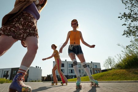 Photo for Long shot of three young active girlfriends on roller skates moving in front of camera along street while spending time in park on sunny day - Royalty Free Image