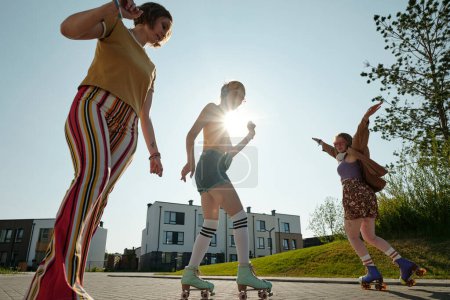 Téléchargez les photos : Trois filles insouciantes en tenue décontractée et patins à roulettes profiter du passe-temps le week-end d'été tandis que l'un d'eux debout contre le soleil brillant - en image libre de droit