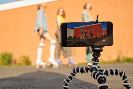 Téléchargez les photos : Focus sur le trépied avec smartphone lors du tournage vidéo de trois jeunes patineuses amicales en tenue décontractée posant contre un mur brun - en image libre de droit