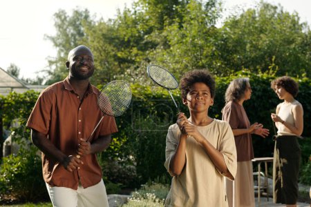 Téléchargez les photos : Heureux jeune homme noir et son fils adolescent avec des raquettes de badminton debout devant la caméra dans un environnement naturel contre deux femmes - en image libre de droit