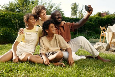 Téléchargez les photos : Heureuse jeune famille interculturelle de quatre personnes prenant des selfies dans un environnement naturel assis sur une pelouse verte devant la caméra - en image libre de droit