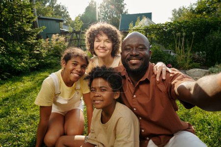 Foto de Selfie retrato de la feliz familia joven intercultural de cuatro mirando a la cámara con sonrisas mientras está sentado en el césped verde en el entorno natural - Imagen libre de derechos