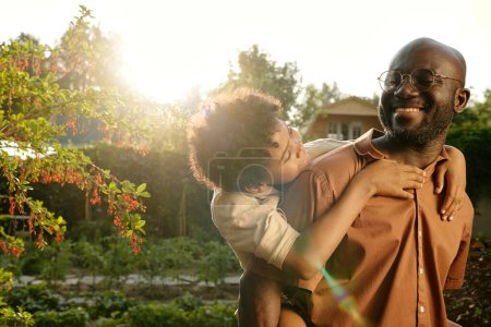 Photo for Young cheerful African American man having fun with his cute son while holding him on back during stroll in the garden on sunny day - Royalty Free Image