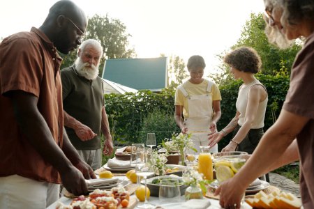 Téléchargez les photos : Grande famille interculturelle heureuse servant une table avec de la nourriture et des boissons faites maison pour le dîner en plein air tout en se tenant contre des buissons verts - en image libre de droit