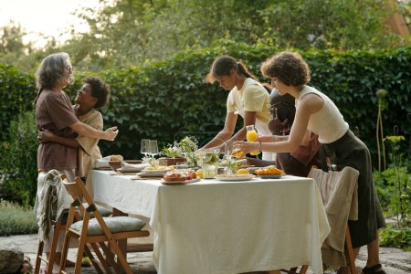 Photo for Cute girl helping her mother serve table with homemade food for outdoor family dinner while happy boy embracing his grandmother - Royalty Free Image