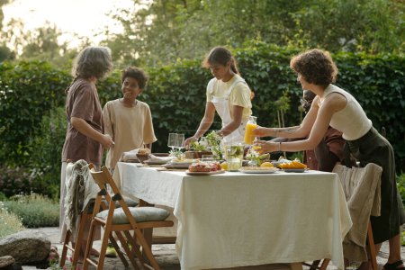 Foto de Uno de los dos hermanos afroamericanos que ayudan a servir mesa para la cena al aire libre en el jardín mientras otro habla con su abuela - Imagen libre de derechos
