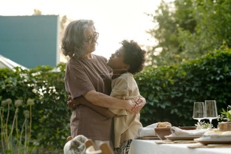Foto de Lindo chico cariñoso en camiseta abrazando a su abuela y mirándola mientras ambos de pie contra la mesa servida antes de la cena familiar - Imagen libre de derechos