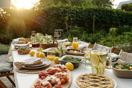 Téléchargez les photos : Partie d'une grande table à manger servie avec divers plats faits maison appétissants et boissons rafraîchissantes debout contre les arbres verts et les buissons - en image libre de droit