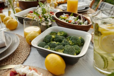 Foto de Contenedor con brócoli al vapor o fresco rodeado de limones y otros alimentos de pie en la mesa servida entre variedad de cosas caseras - Imagen libre de derechos
