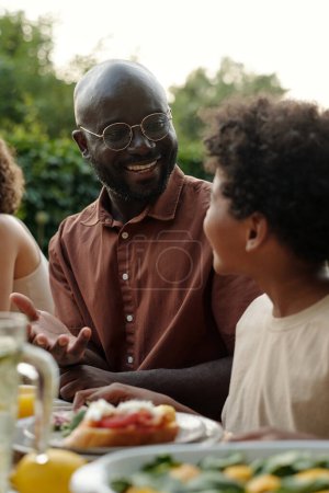 Foto de Feliz joven afroamericano hombre en gafas charlando con su lindo hijo mientras está sentado junto a él por la mesa servida durante la cena familiar al aire libre - Imagen libre de derechos