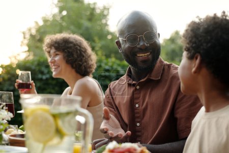 Téléchargez les photos : Jeune homme afro-américain souriant regardant son fils mignon pendant la communication tandis que les deux assis à table servie pendant le dîner en plein air - en image libre de droit