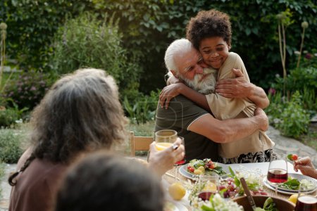 Photo for Affectionate senior man embracing his African American grandson while sitting by dinner table served with healthy and tasty homemade food - Royalty Free Image