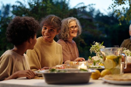 Foto de Linda chica afroamericana mirando a su hermano durante la comunicación mientras ambos sentados junto a la mesa servida durante la cena familiar al aire libre - Imagen libre de derechos