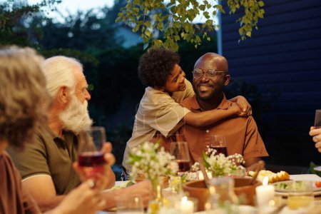 Téléchargez les photos : Joyeux garçon mignon embrassant son jeune père souriant et le regardant pendant le dîner de famille en plein air avec les grands-parents assis à table - en image libre de droit